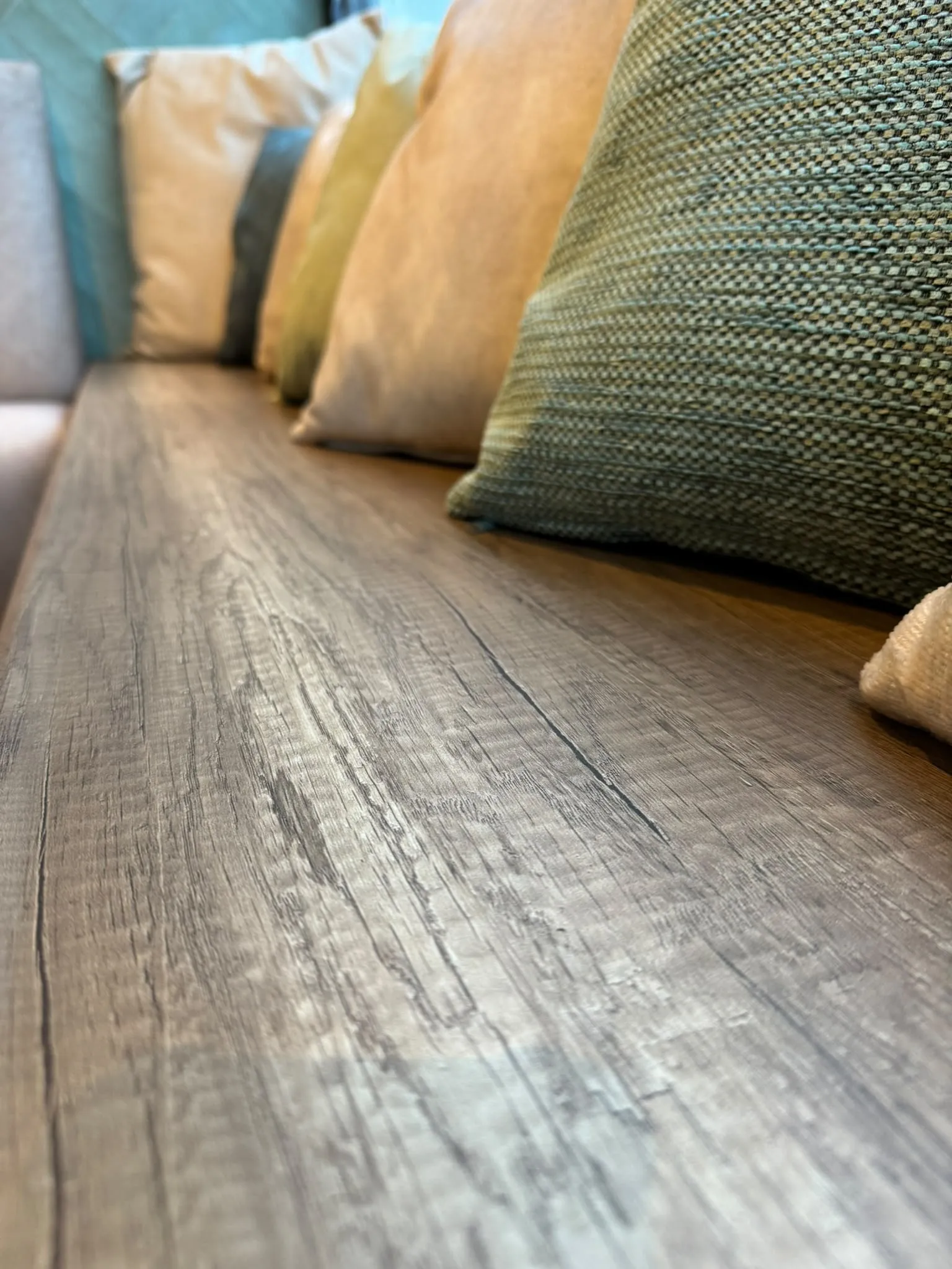 Detail van een houten tafelblad met daarachter een rij kussens in beige, oker en zeegroen
