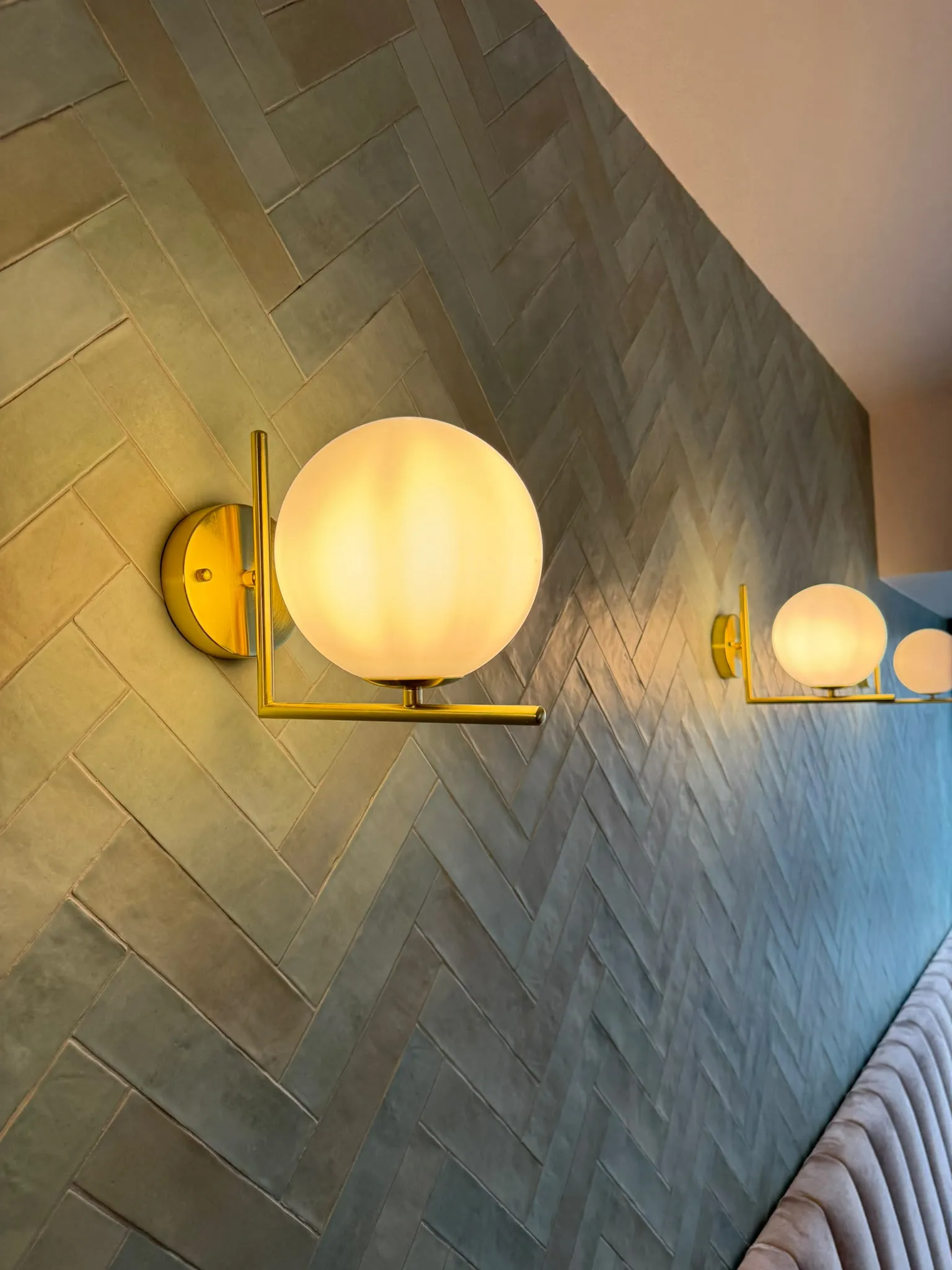 Detail van een gouden wandlamp met melkglazen bol tegen de handgemaakte zeegroene zellige-tegels in visgraatverband