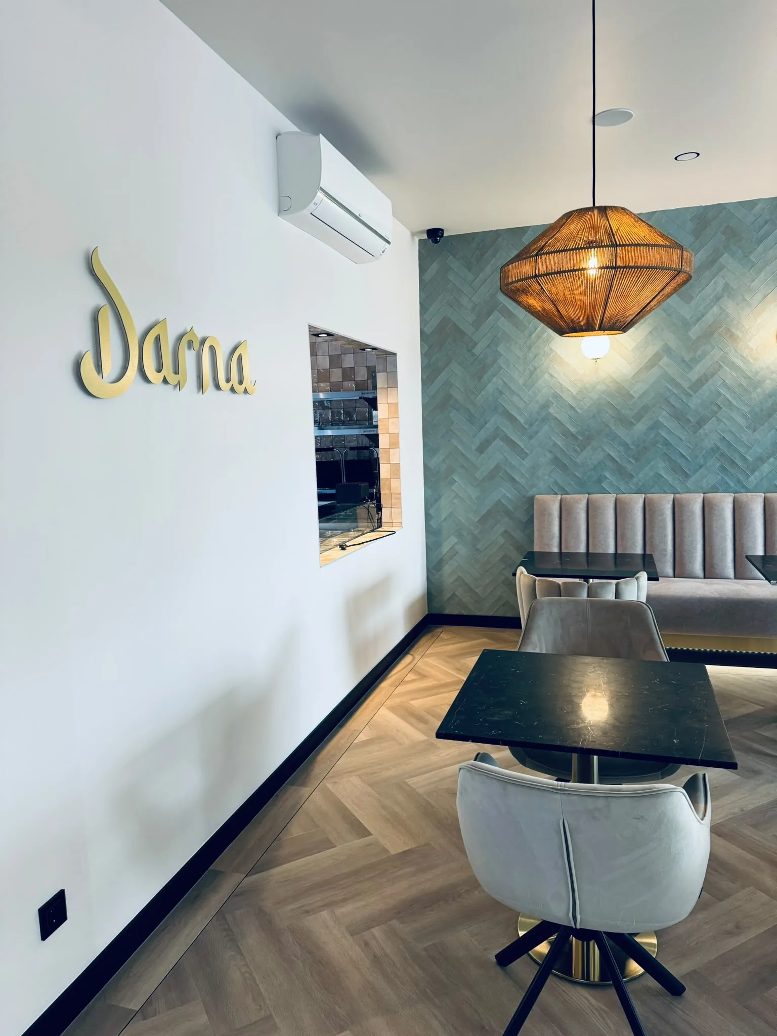 De witte wand met het gouden Darna-logo in schrijfletters, met daarnaast een doorgeefluik naar de keuken en een rieten hanglamp boven een marmeren tafel