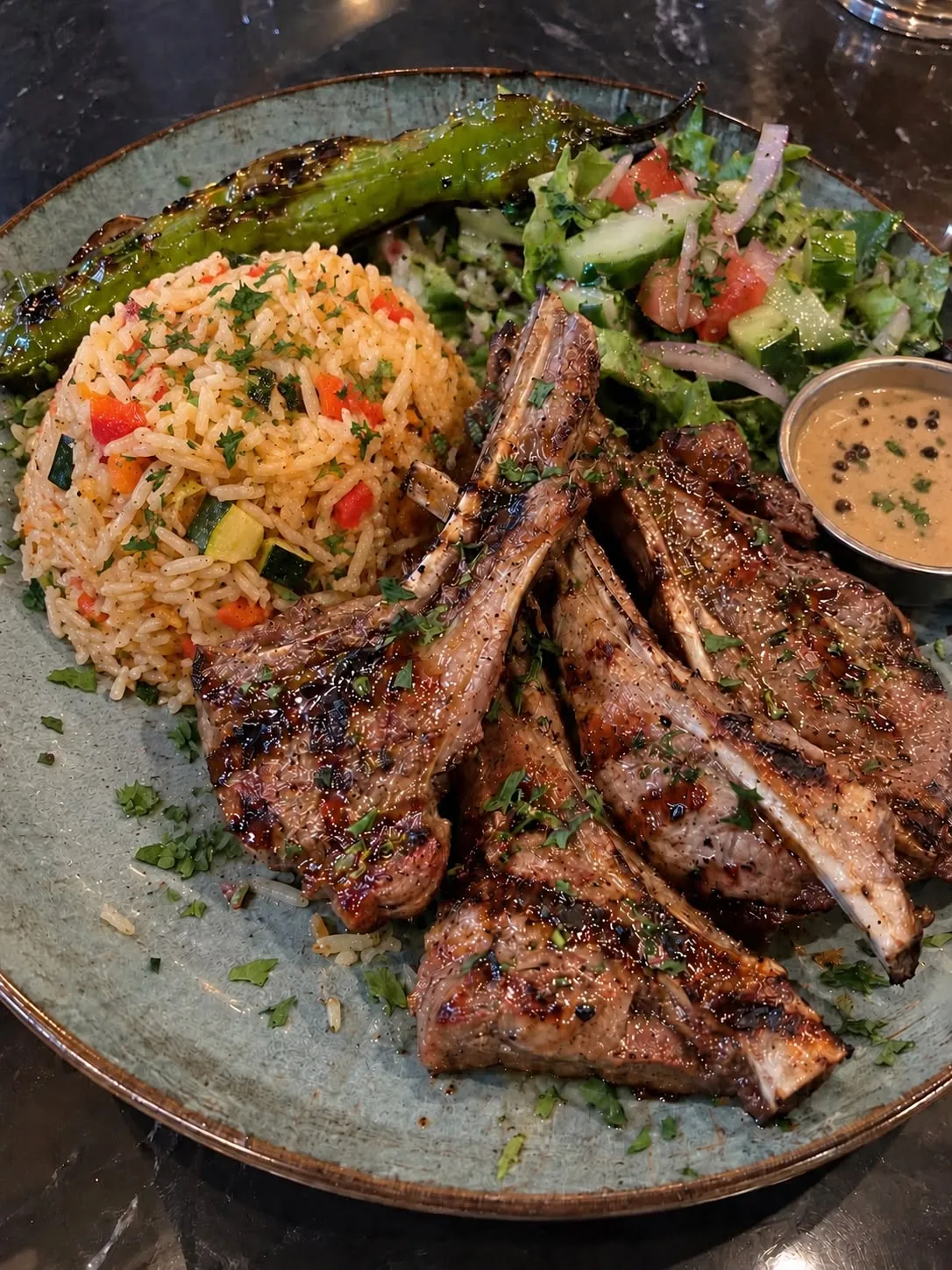 Een bord lamskoteletten bij Darna: gegrilde koteletjes met peterselie, kruidige rijst met paprika en courgette, een frisse salade van tomaat en komkommer, een gegrilde groene peper en een kommetje pepersaus