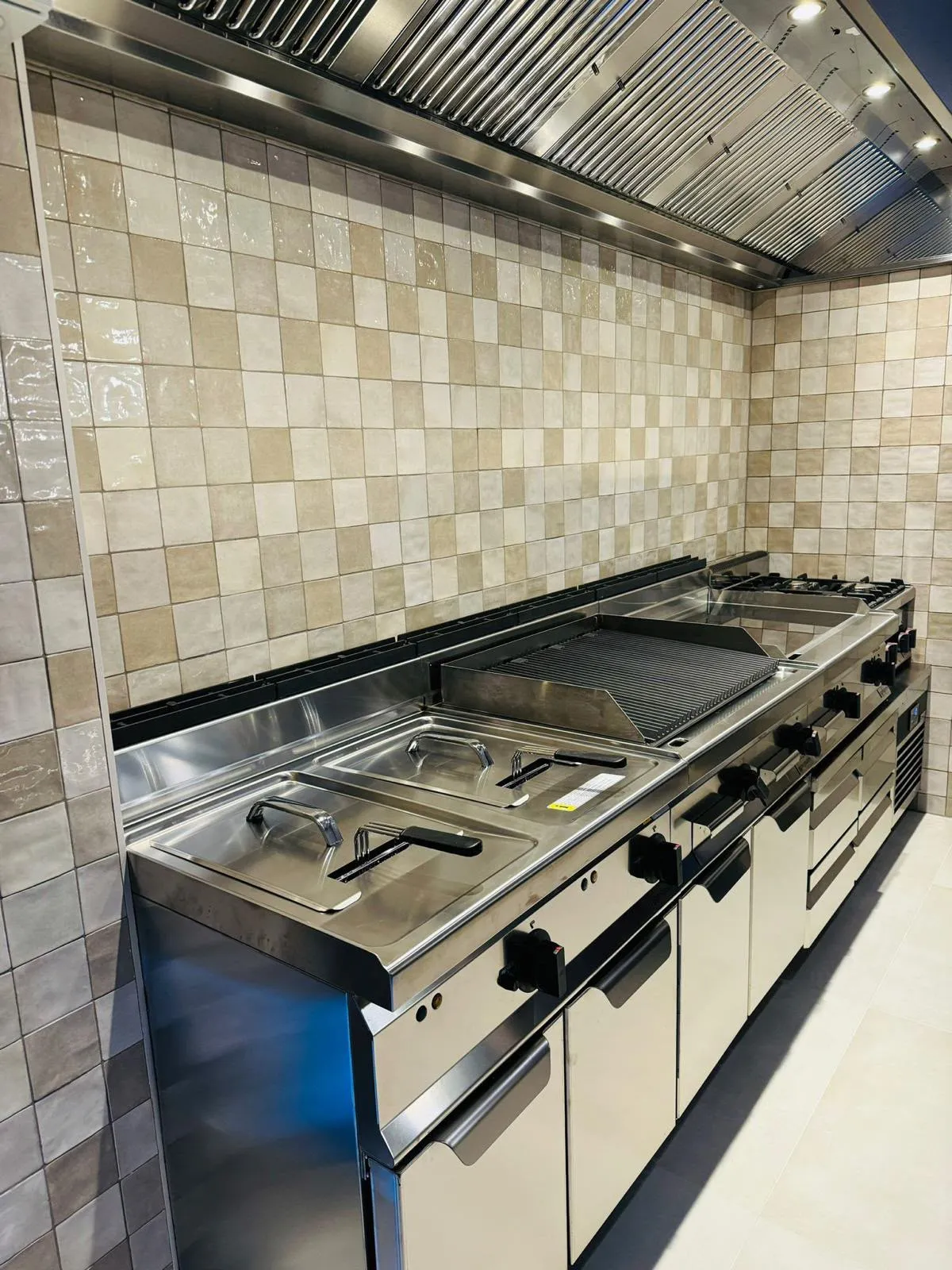 De keuken van Darna: in het midden de lavagrill met zijn gietijzeren rooster, links twee frituurpannen en rechts gaspitten, onder een afzuigkap van rvs tegen een wand van lichte tegels