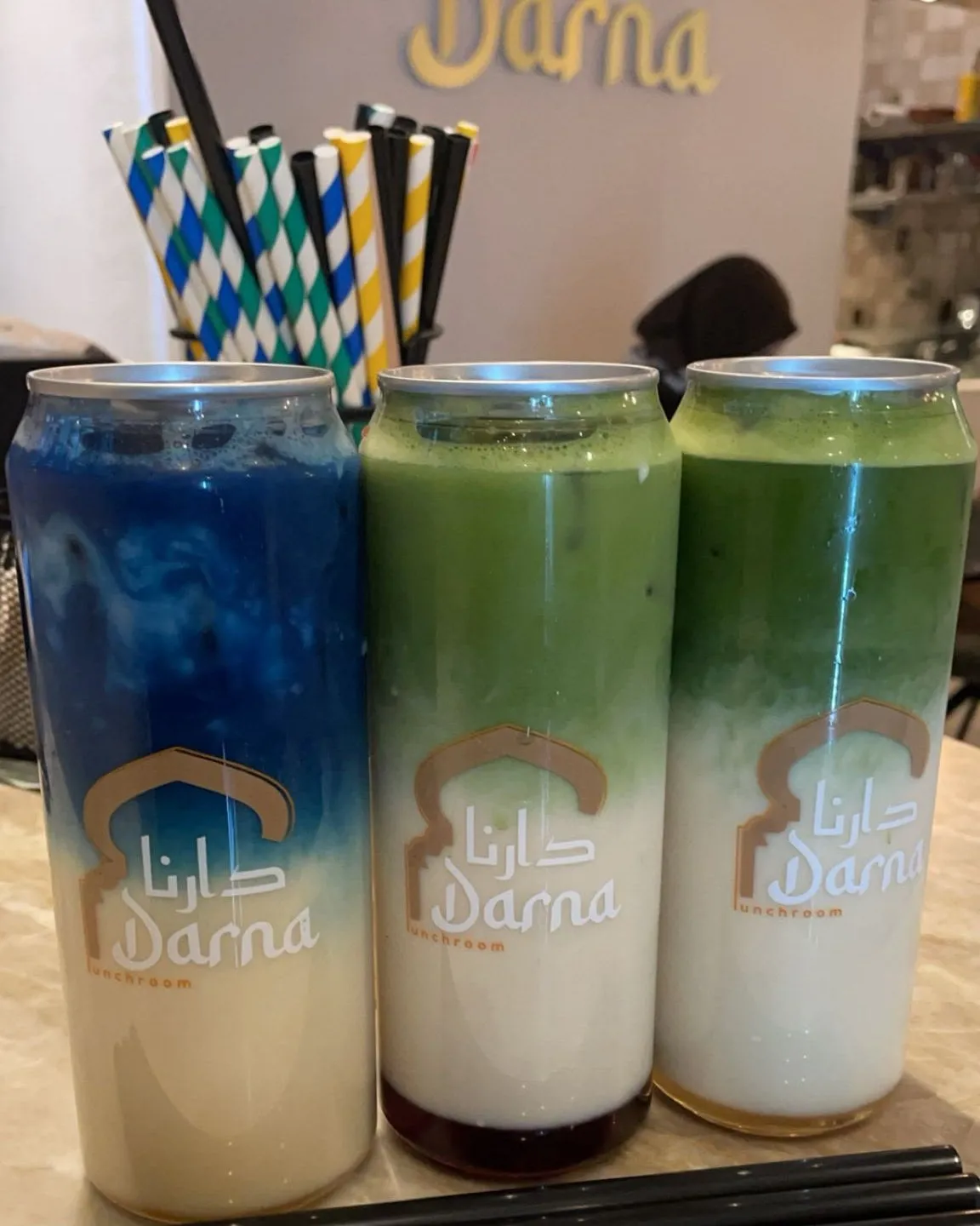 Drie blikjes matcha op de bar bij Darna: links de blauwe Darna matcha special, rechts twee groene matcha's, met de melk onderin en het gouden Darna-logo op de muur erachter