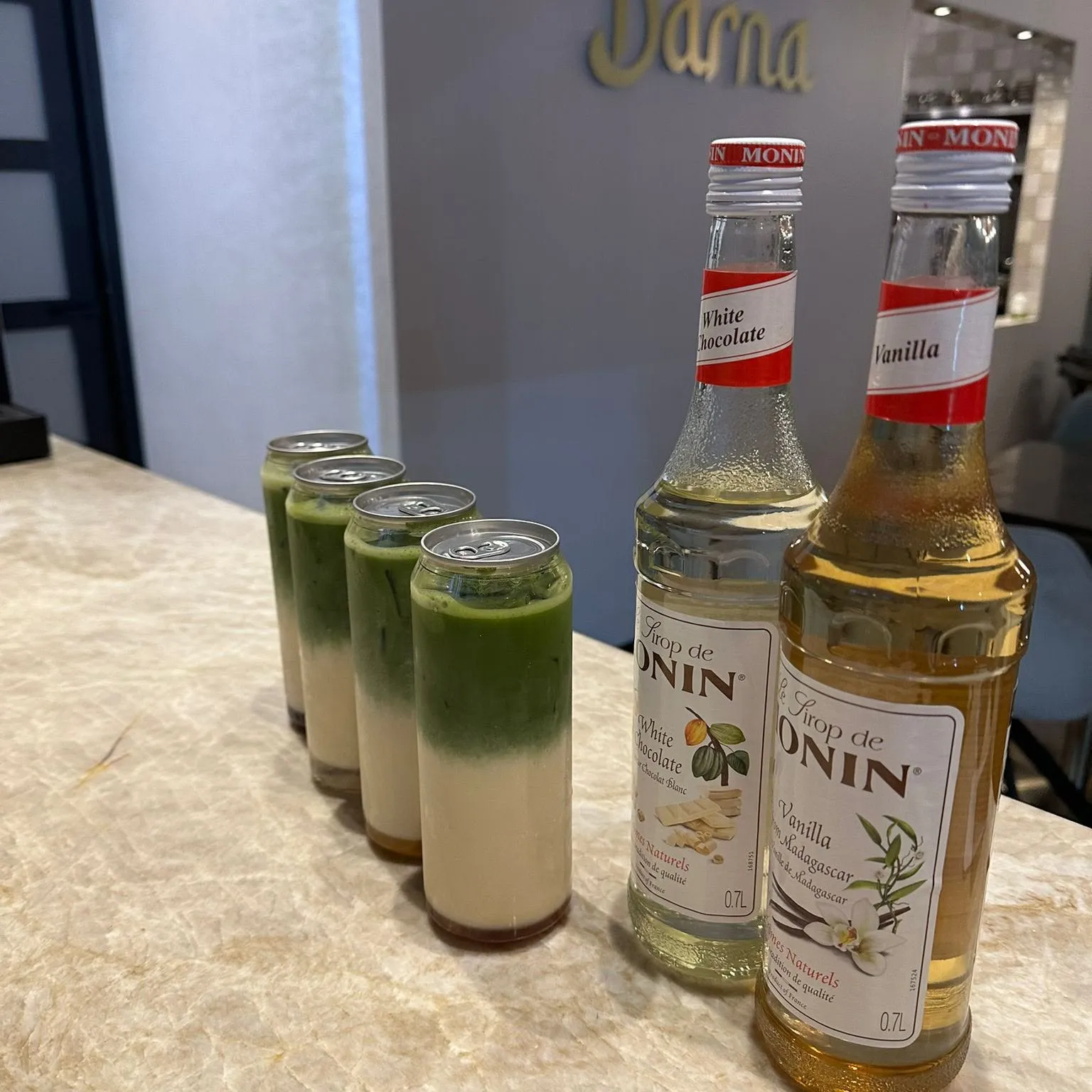 Vier blikjes groene matcha op een rij op de marmeren bar, naast flessen Monin-siroop in de smaken witte chocolade en vanille