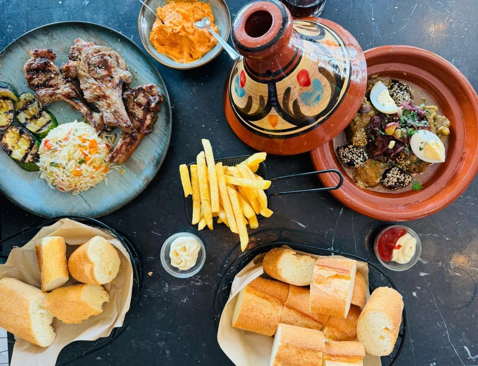 Een gedekte tafel bij Darna van bovenaf: lamskoteletten van de lavagrill met rijst en gegrilde courgette, een beschilderde tajine, zaalouk met ei en olijven, friet en manden vers brood